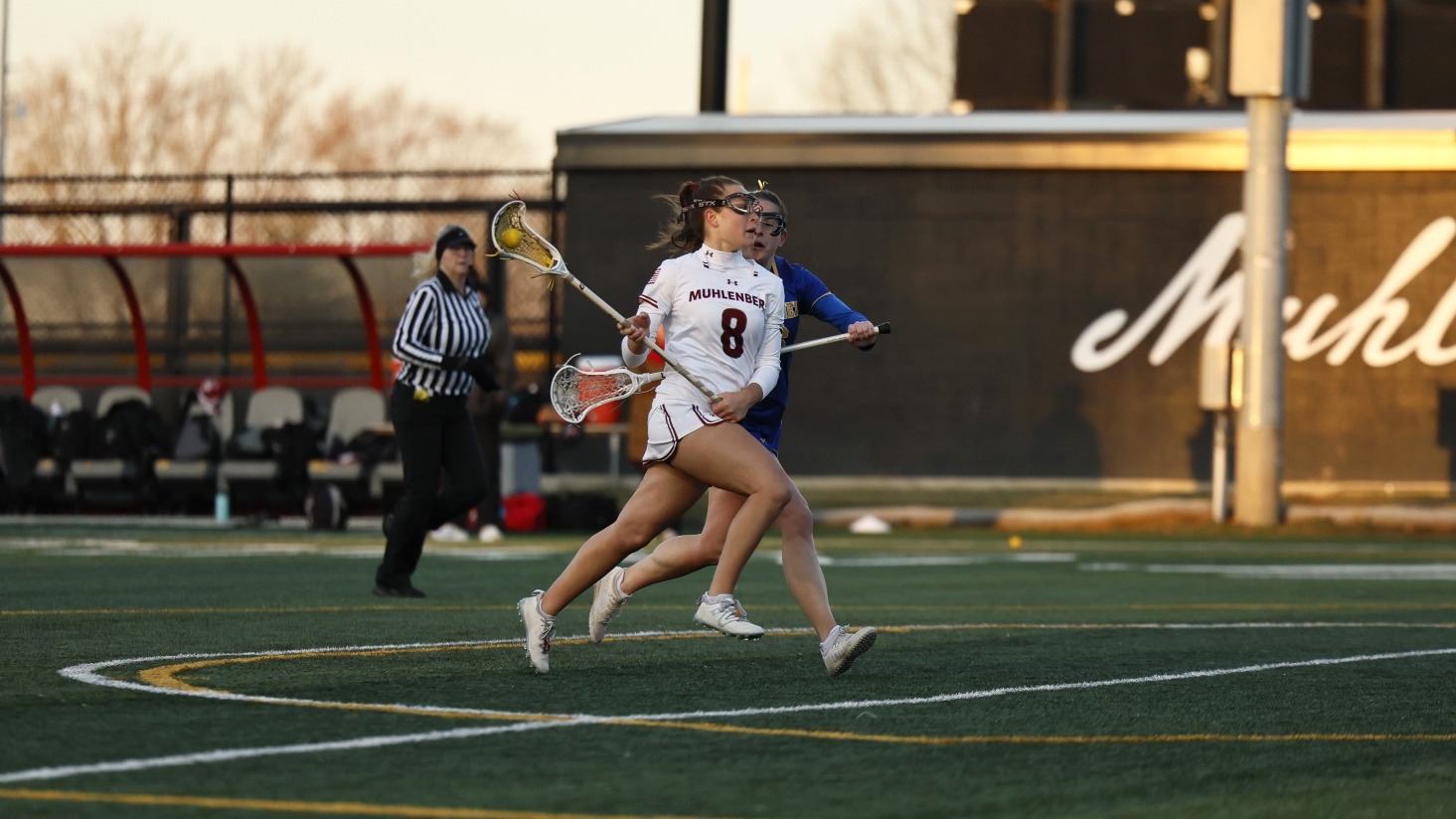 A Muhlenberg women’s lacrosse player wearing number 8 sprints down the field while closely defended by an opponent during an evening game.