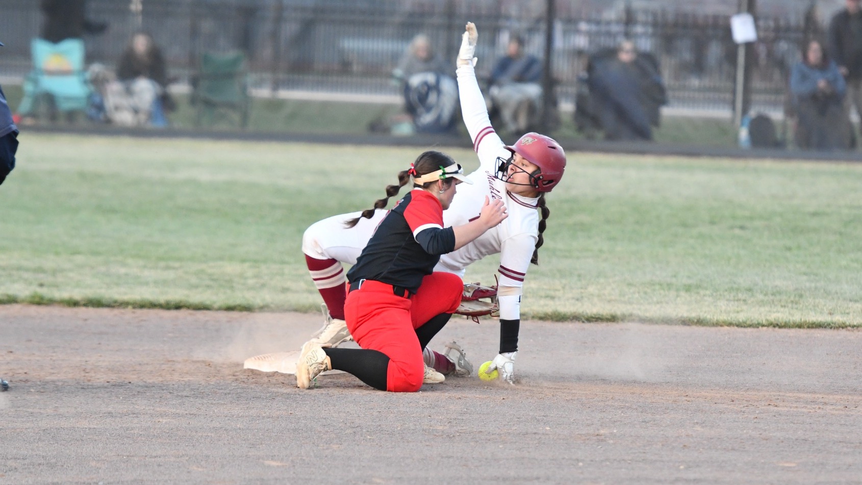 A softball runner in a white uniform slides into a base as a fielder in red pants applies a tag, kicking up dirt.