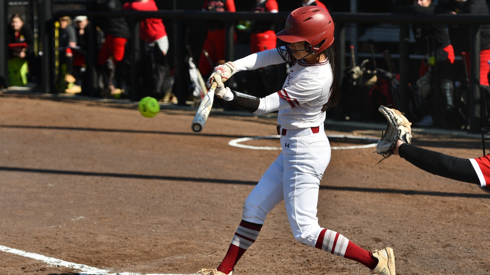 A softball player in a white uniform and red helmet swings and makes contact with the softball while a catcher reaches forward behind her and teammates watch from the dugout.