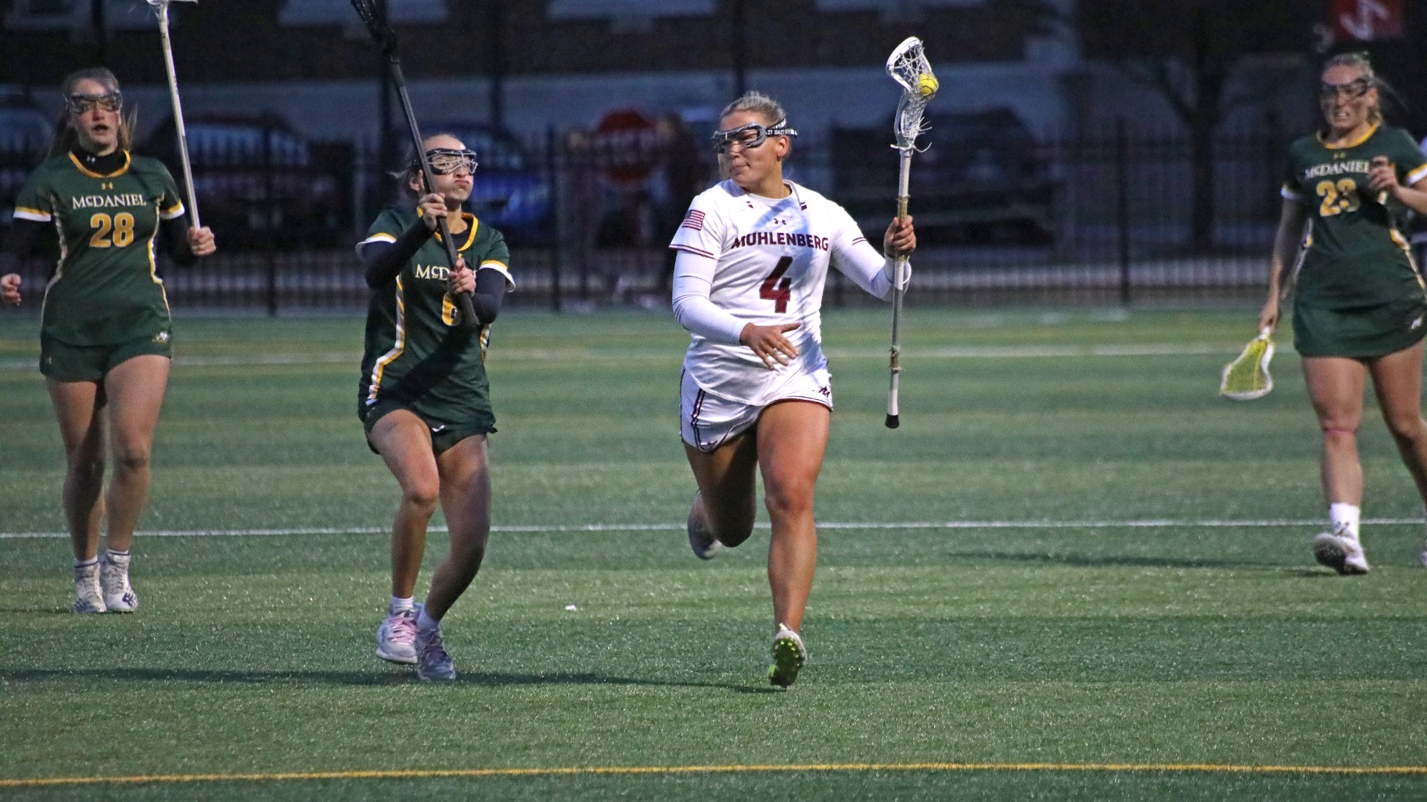 A Muhlenberg player in a white uniform carries the ball upfield while two McDaniel defenders in green chase behind during a women’s lacrosse game.