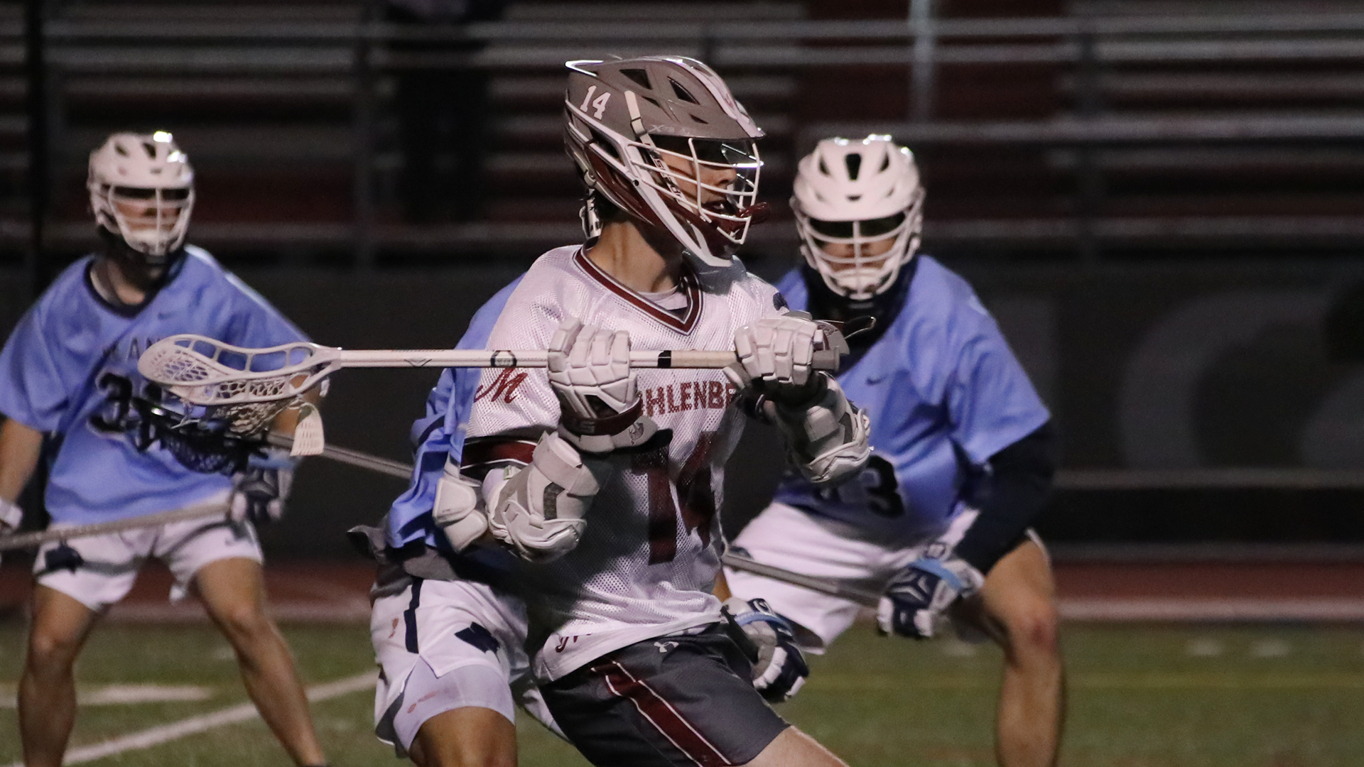 a lacrosse player in a white uniform and grey helmet facing to the right, with players in light blue uniforms behind him