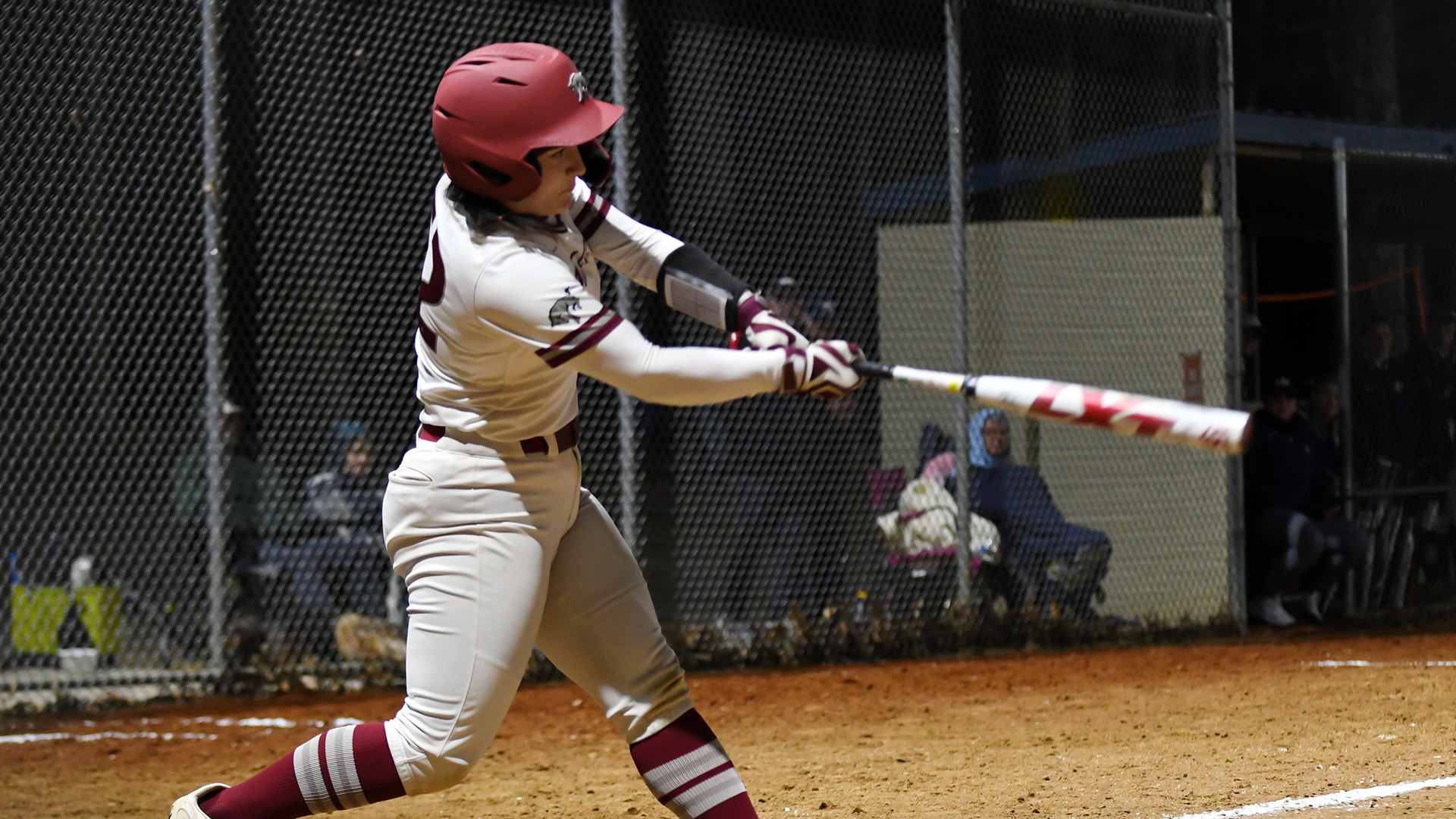 a softball player in a white jersey and red helmet follows through on her swing during a night game