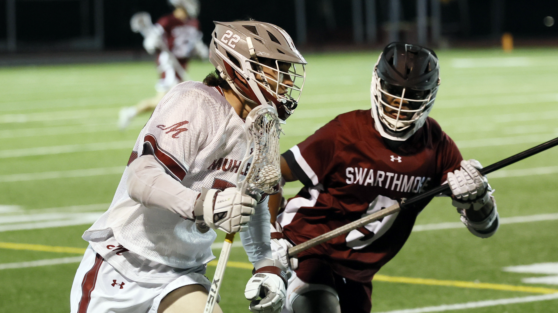 a lacrosse player in a white jersey and grey helmet carries the ball while being guarded by a player in a red jersey and black helmet