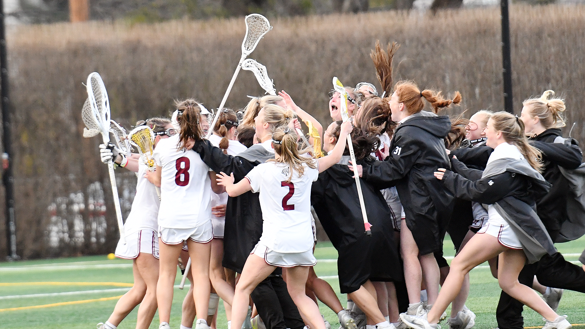 women's lacrosse players in white uniforms and black jackets celebrate a win