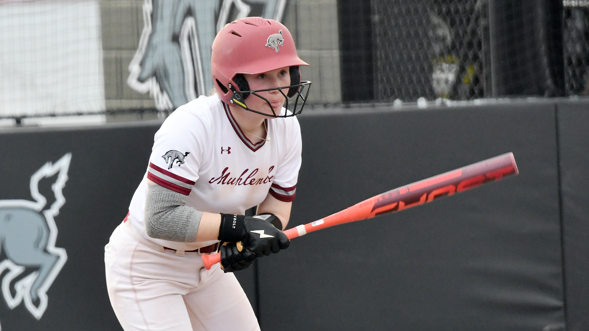 a softball player in a white jersey and red helmet follows the flight of the ball after swinging