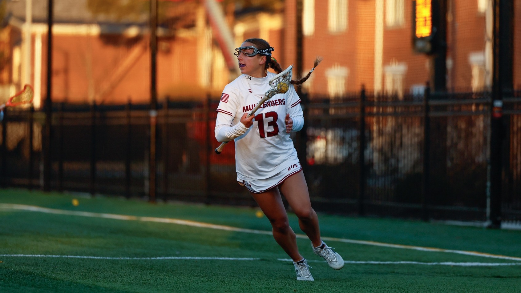 A Muhlenberg women’s lacrosse player wearing #13 in white runs with the ball in her stick during a game.