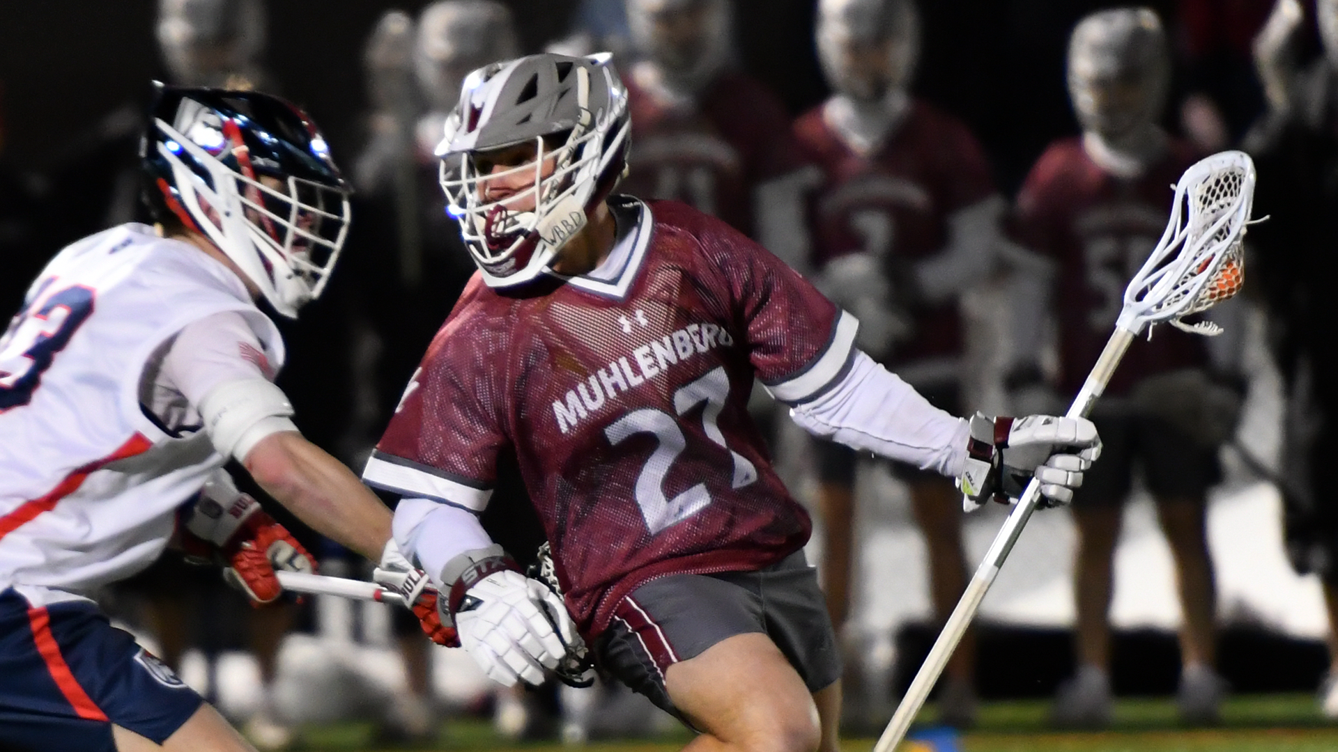a lacrosse player in a red jersey, grey shorts, and a grey helmet holds the ball in his stick away from a defender in a white jersey, blue shorts, and a blue helmet