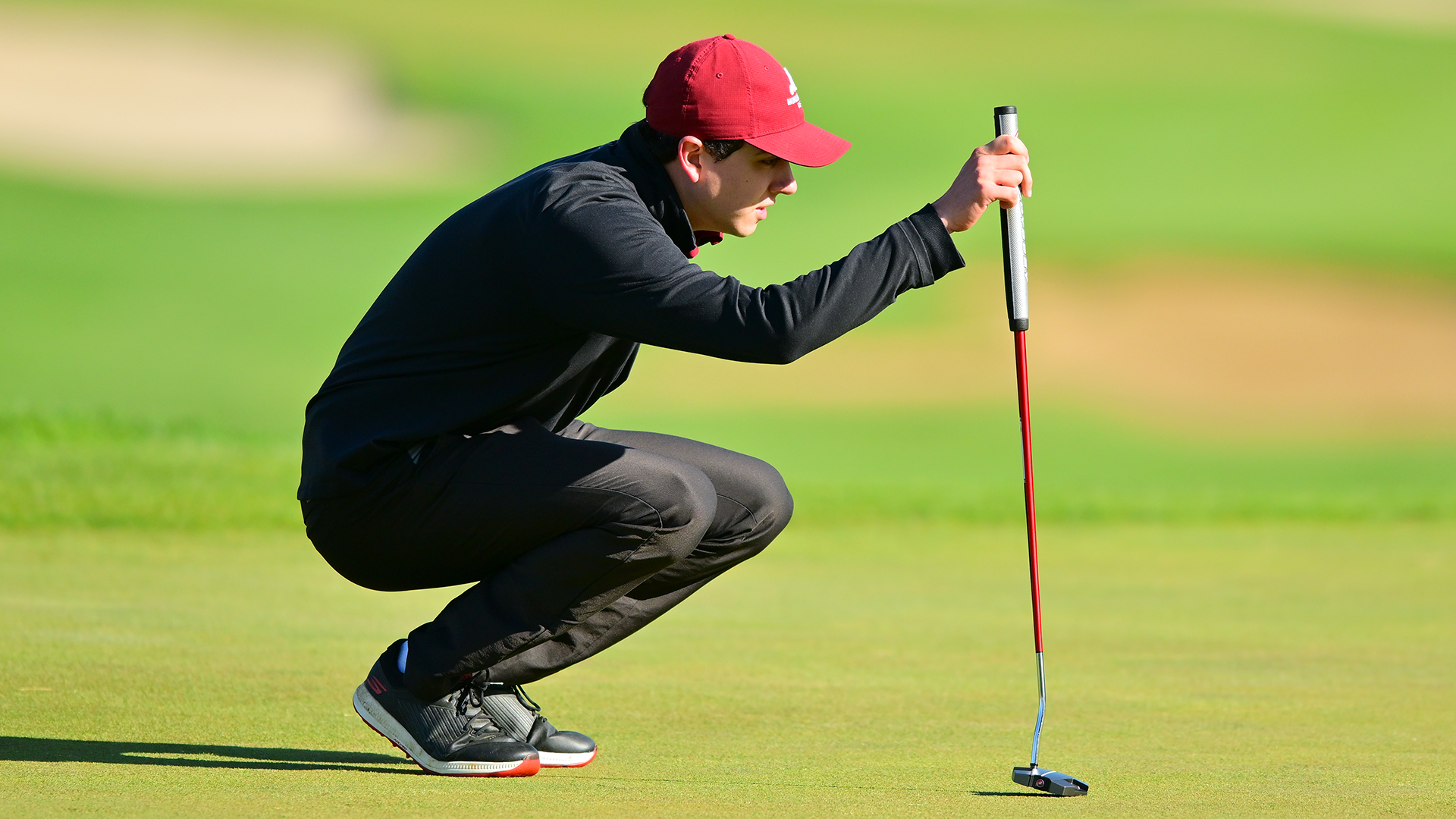 side view of a golfer in a black jacket and black pants squatting on the green to line up a putt