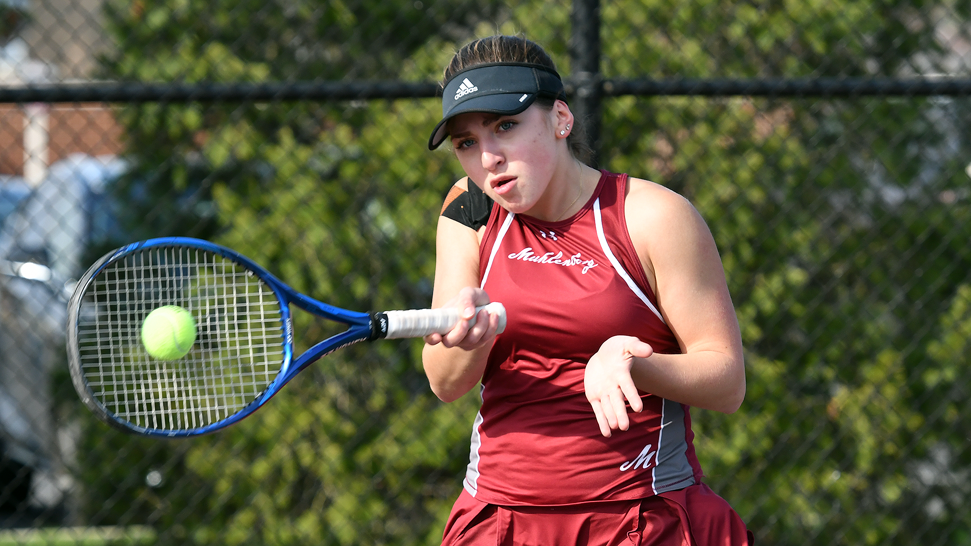 a tennis player in a red sleeveless uniform and black visor connects with the ball on a forehand