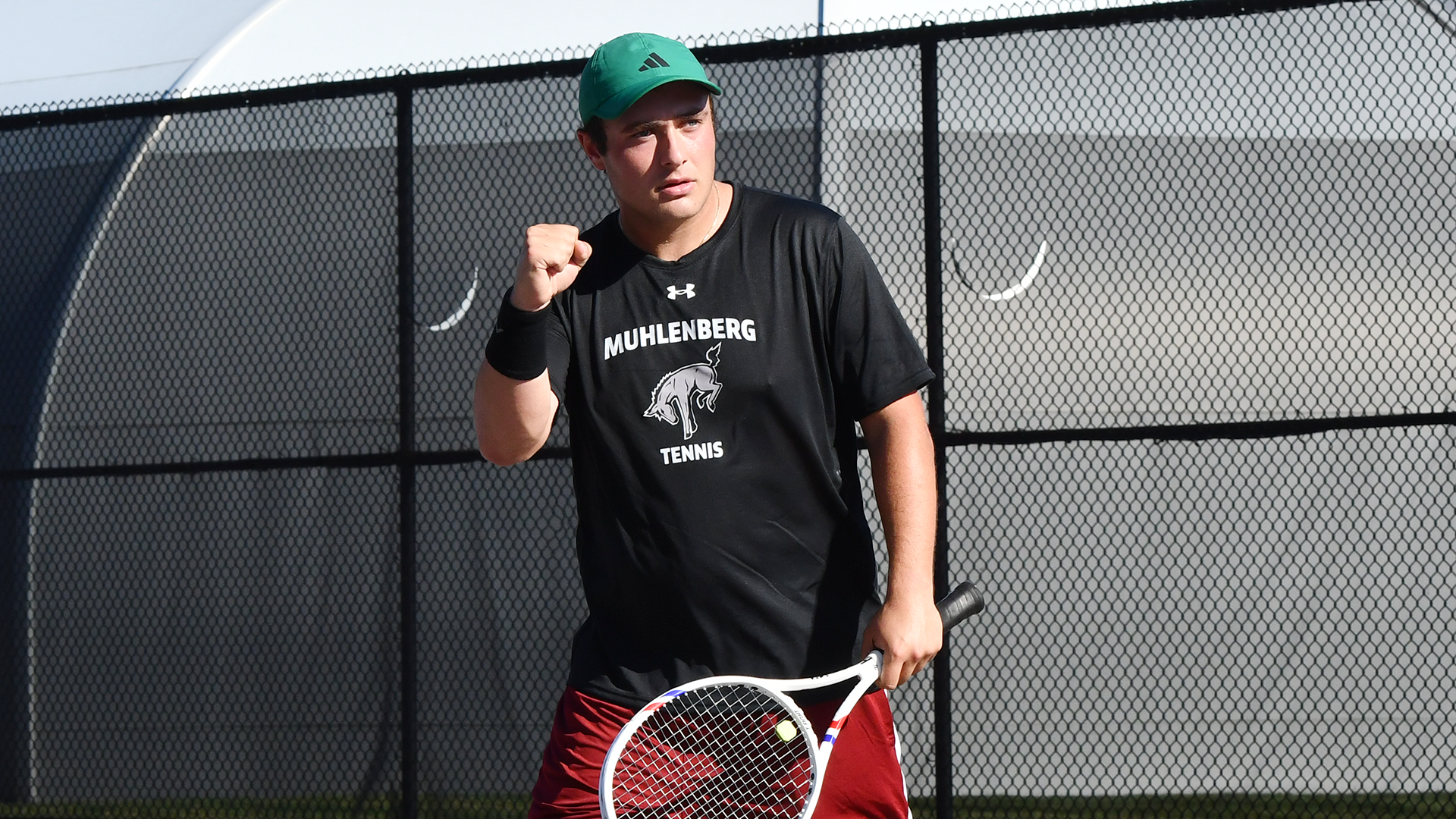 a tennis player in a black t-shirt, red shorts and green hat pumping his fist