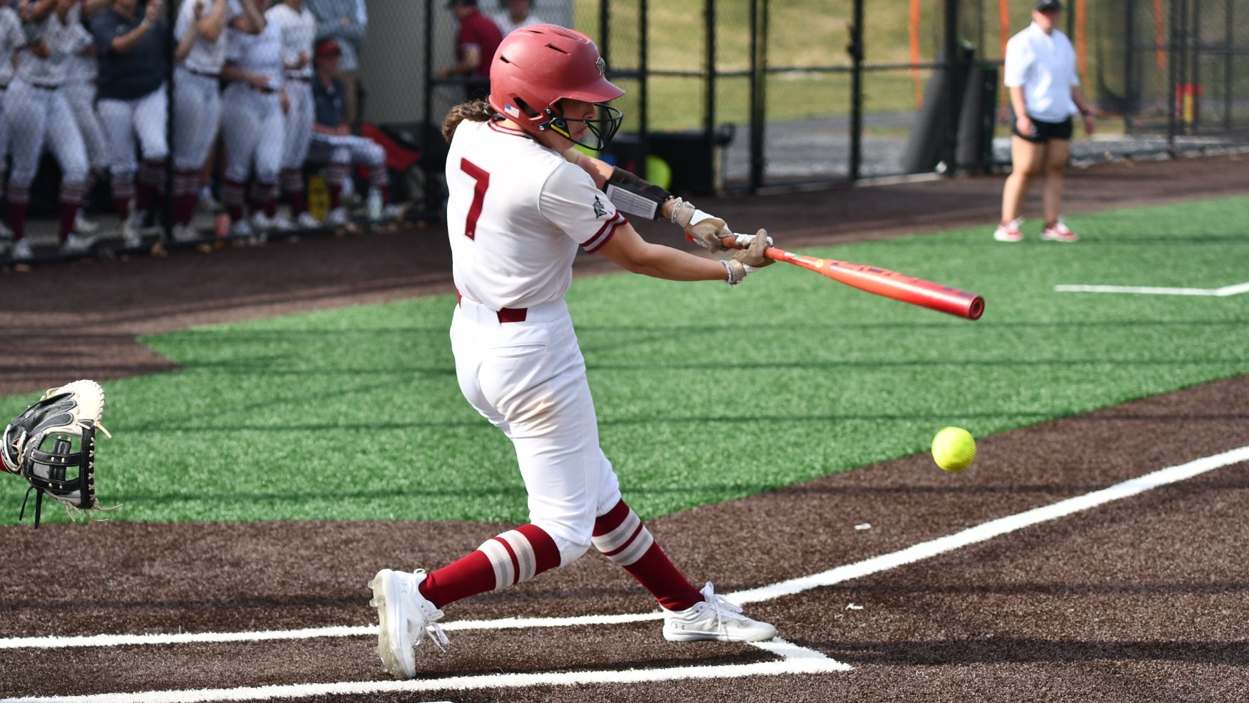 A softball player wearing #7 in white makes contact with a pitch at home plate as teammates watch from the dugout behind her.