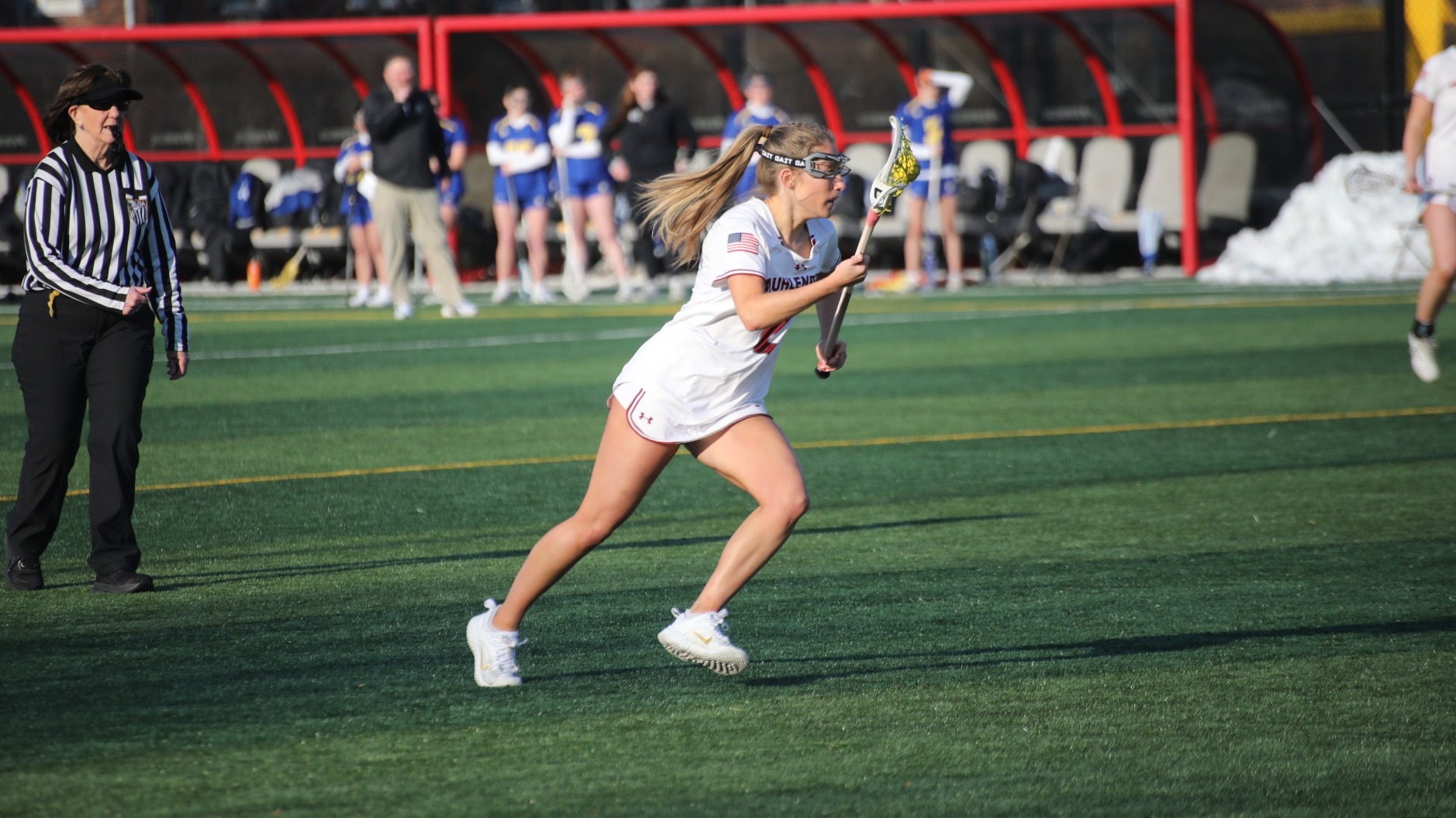 A Muhlenberg women’s lacrosse player in white runs upfield with the ball in her stick during a game while a referee and players on the sideline watch in the background.