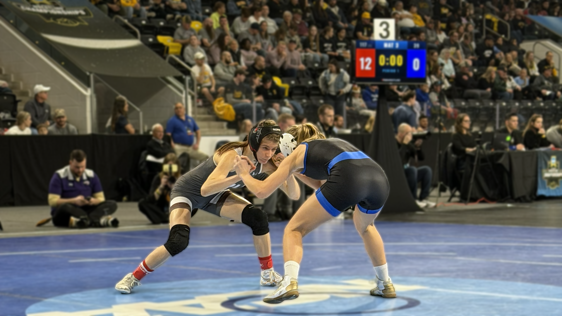 two wrestlers, one in a grey singlet and one in a black singlet with blue stripes, square off on a mat in a large arena