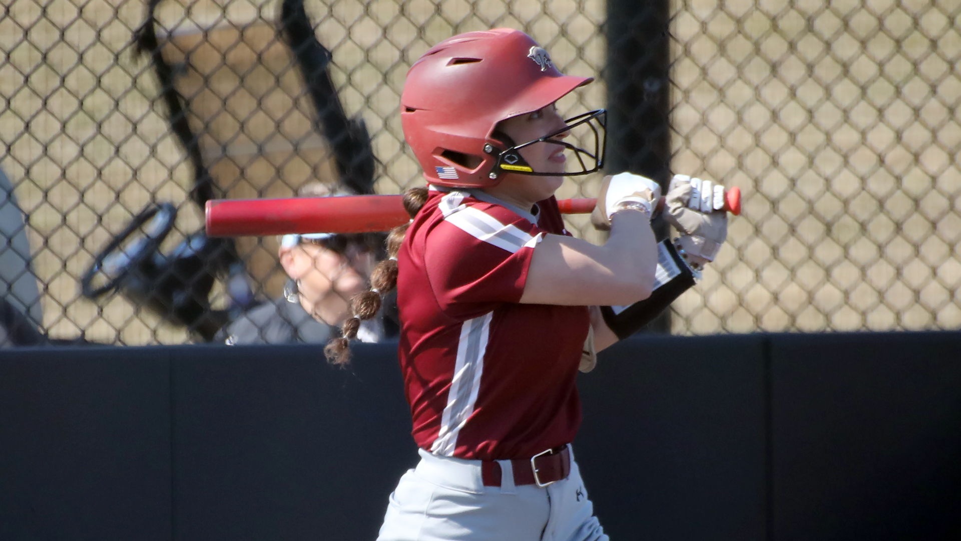 a softball batter in a red jersey and red helmet follows the flight of the ball after swinging