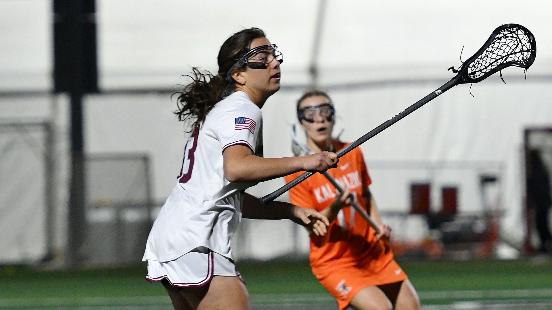 a lacrosse player in a white uniform facing right extends her stick with a player in an orange uniform in the background