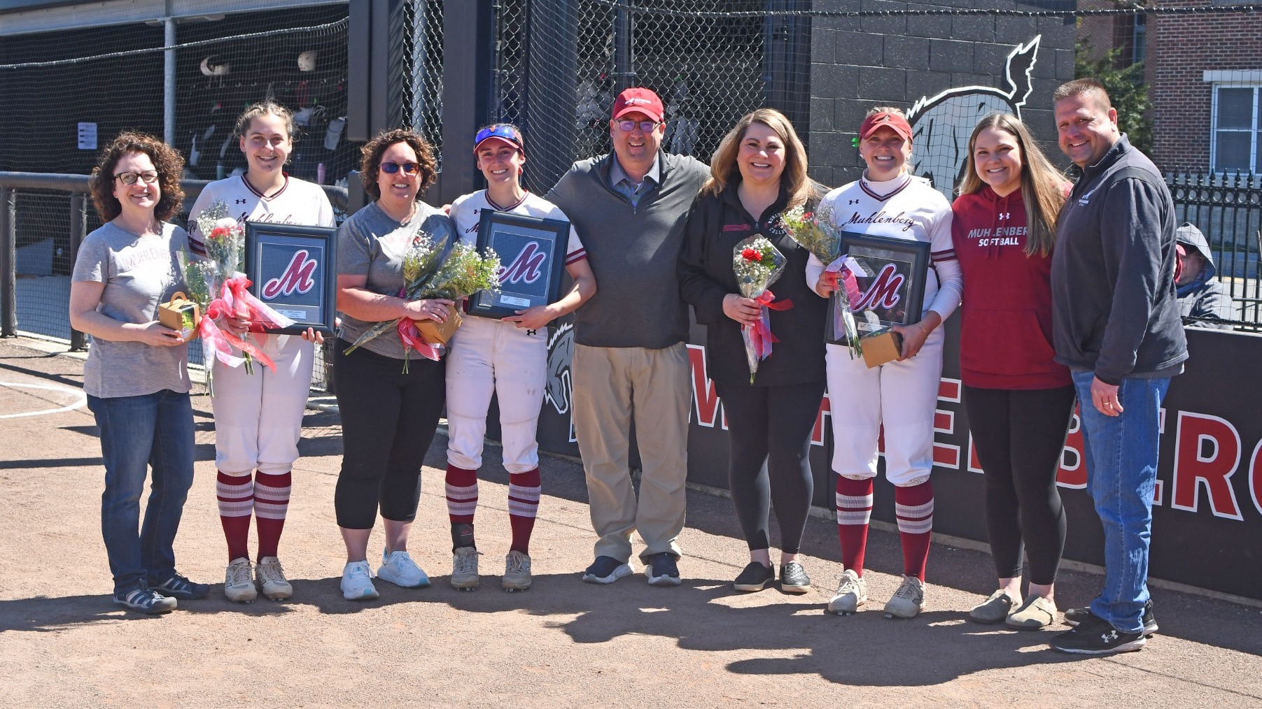 A group of Muhlenberg softball players and their family members pose on the field holding flowers and framed jerseys during the senior day celebration.