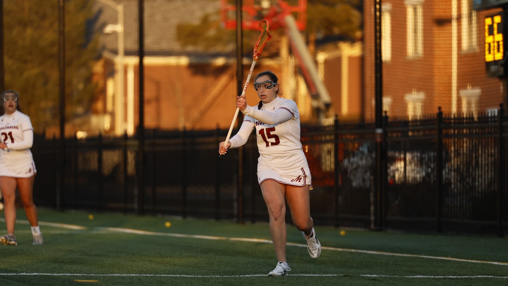 A women’s lacrosse player in a white uniform runs forward while cradling the ball during the afternoon.