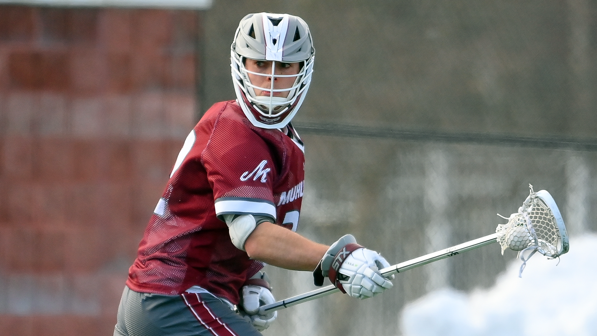 a men's lacrosse player in a red uniform and grey helmet with the ball in his long stick