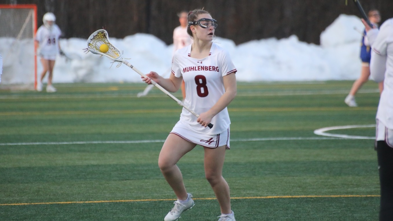 A women's lacrosse player cradles the ball in her stick while wearing white