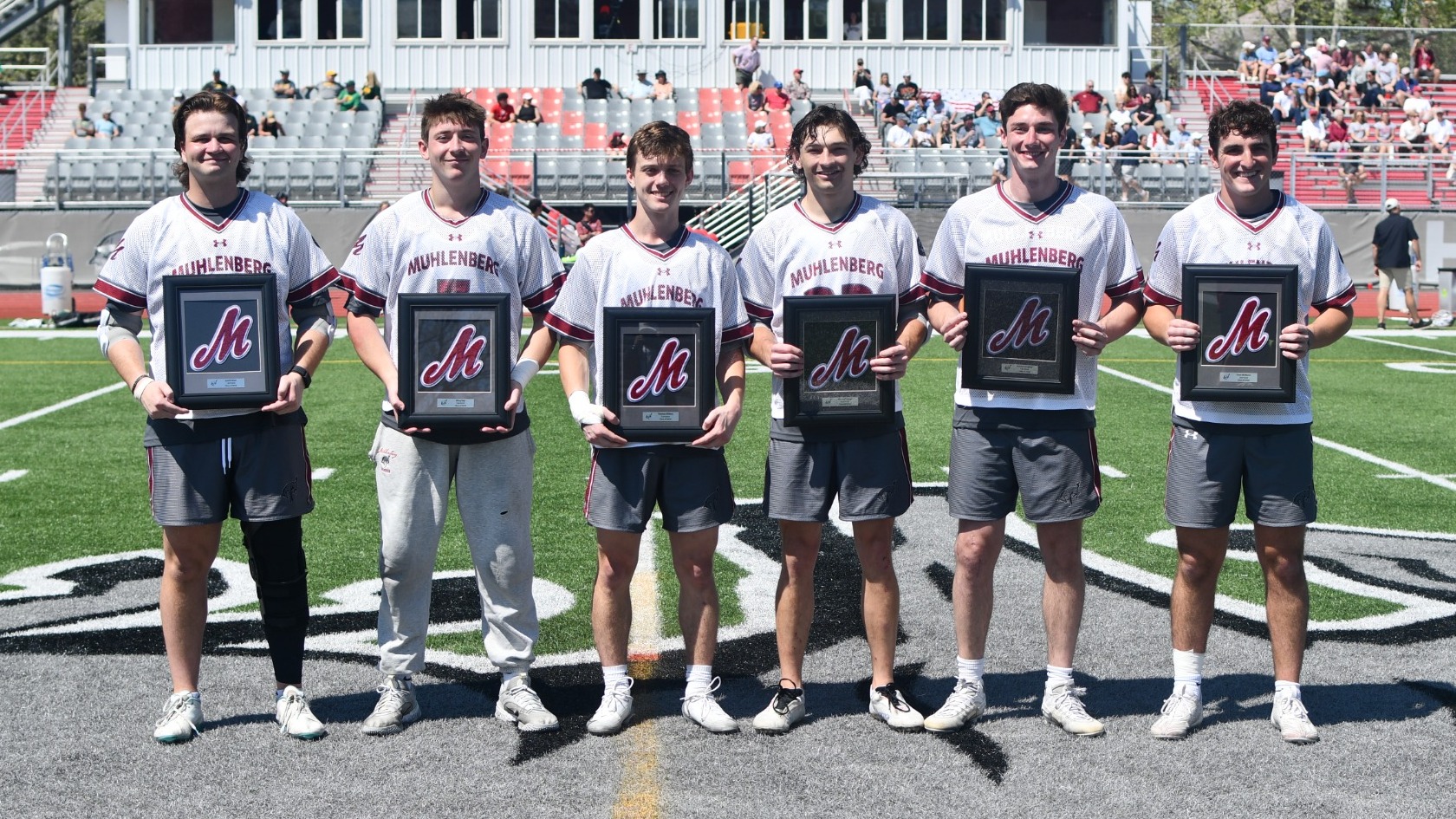 six men's lacrosse players stand and pose with plaques at midfield