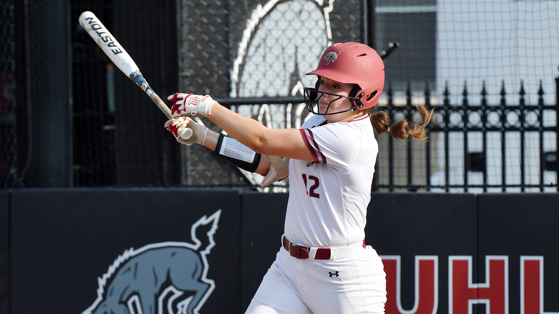 a left-handed softball batter in a white jersey and red helmet following through on her swing