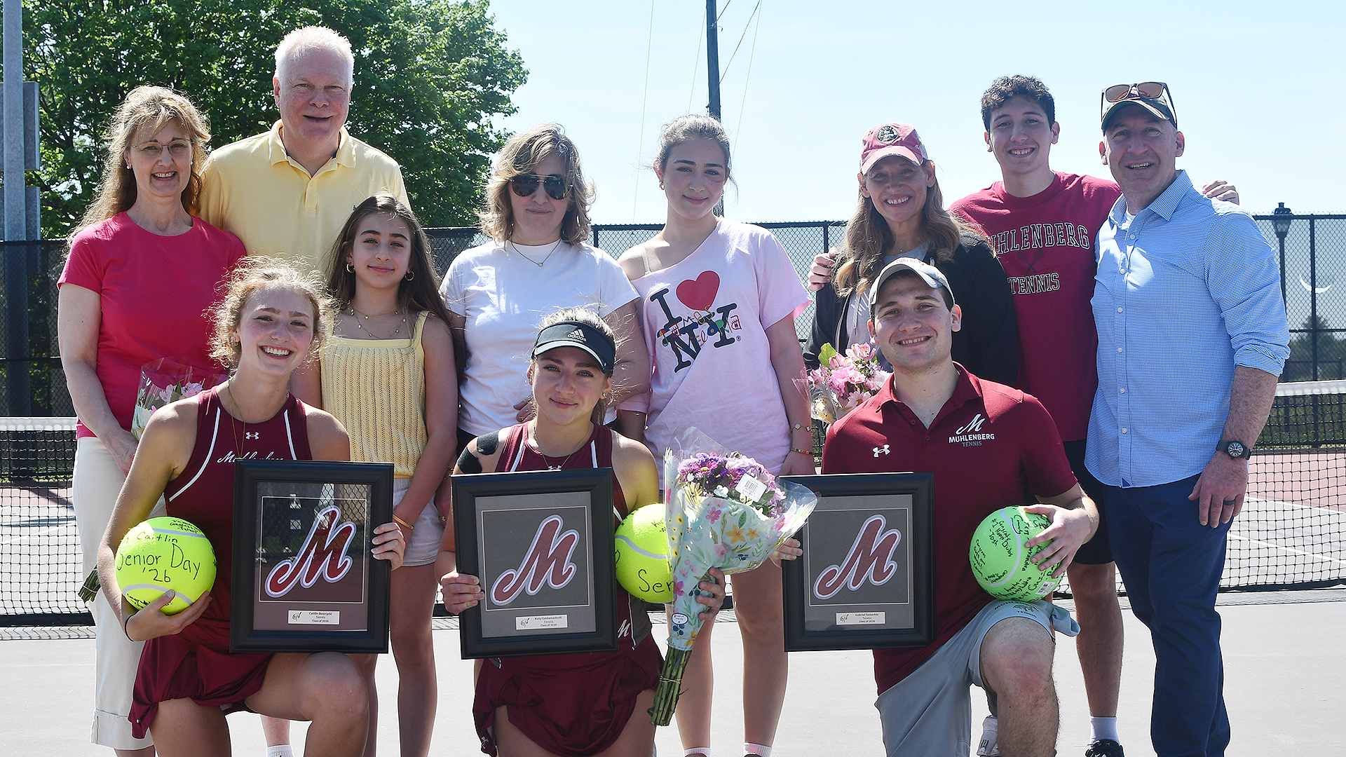 group picture of tennis players holding plaques kneeling on the court with their families standing behind them