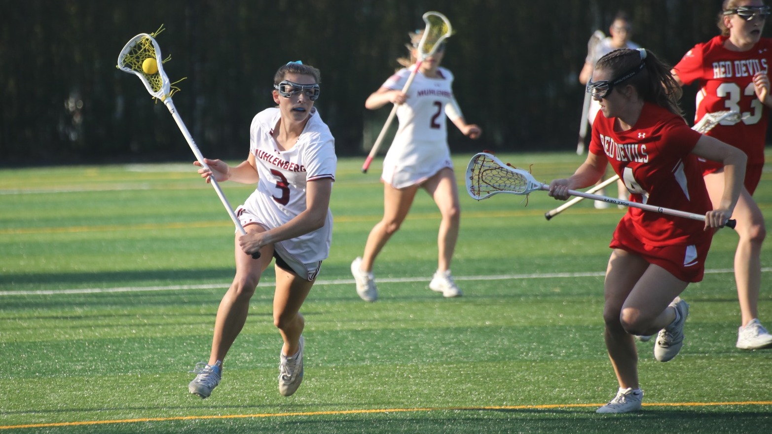 A women's lacrosse player in white carries the ball by a defender in red.