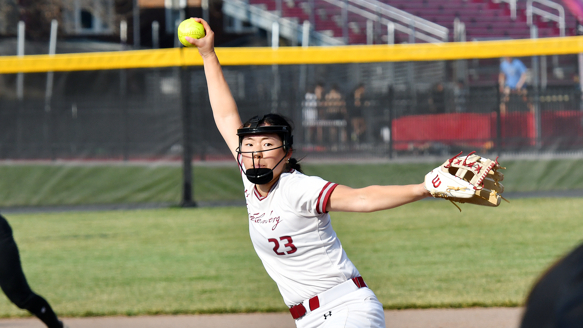 a softball pitcher in a white uniform delivering a pitch on a sunny day