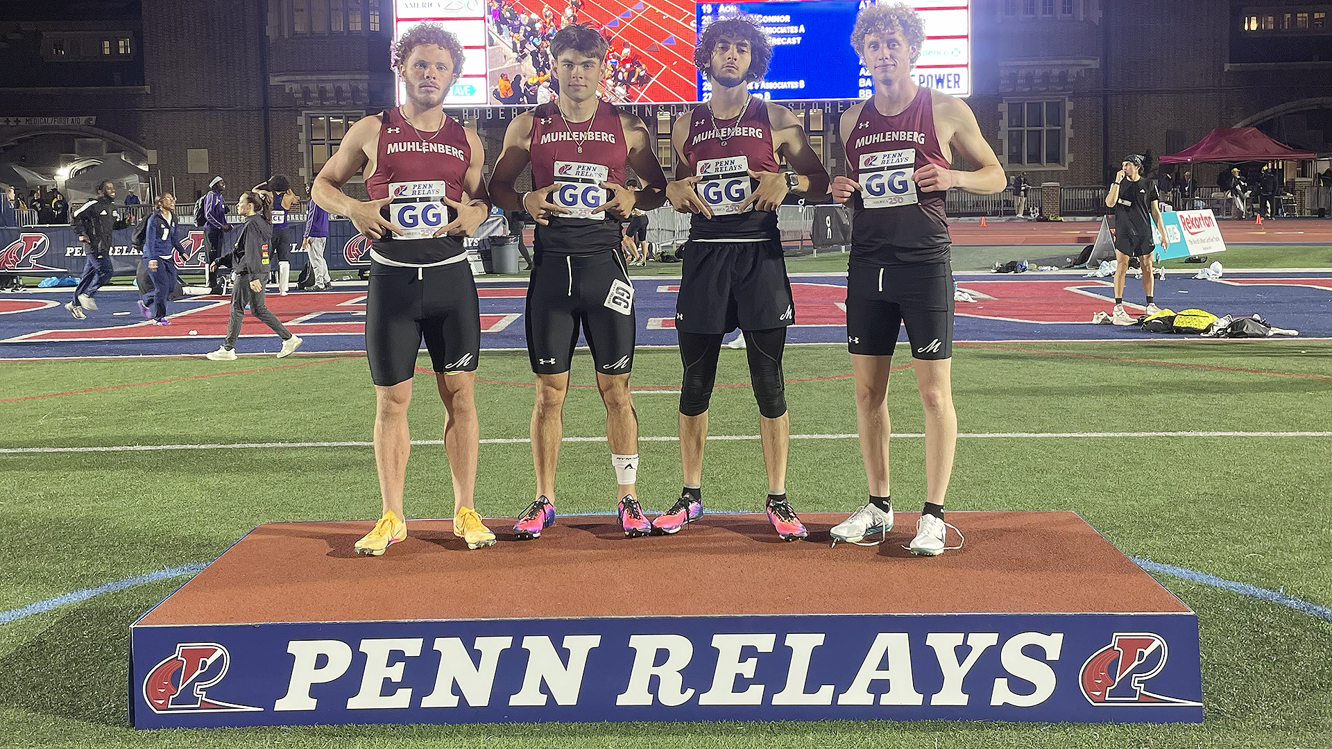 four runners in red tops and black shorts standing on a podium with a video board behind them
