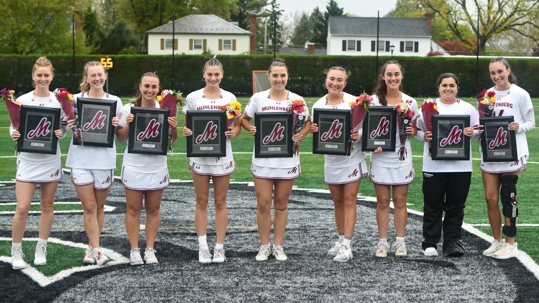 8 women's lacrosse players in white line up and pose for a photo while holding flowers and a plaque