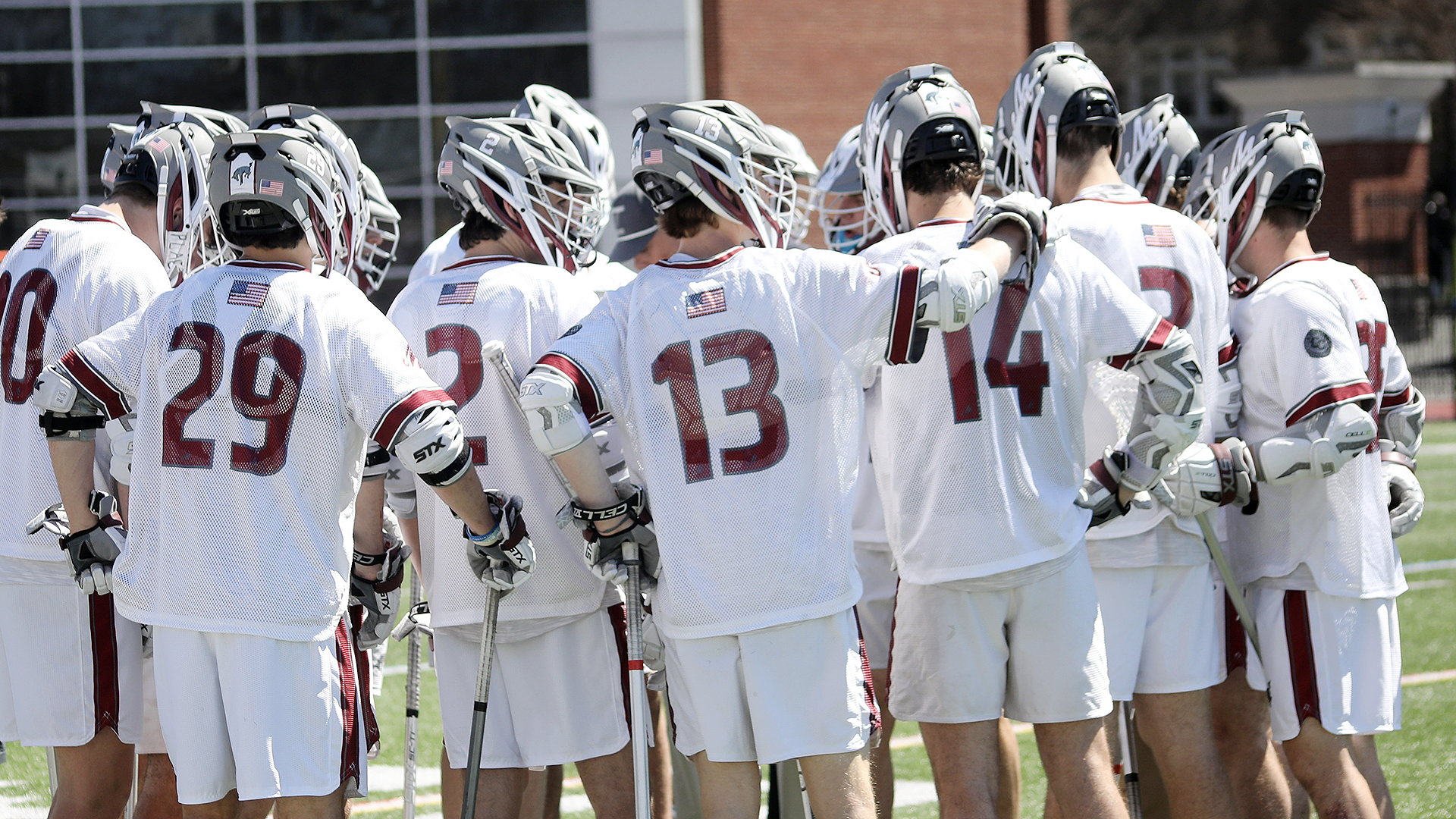 men's lacrosse players in white jerseys and grey helmet standing in a huddle outdoors on a sunny day