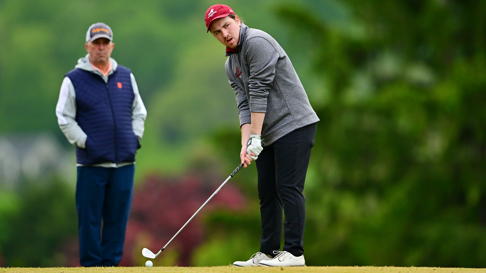 a golfer in a red hat, grey pullover, and black pants getting ready to hit with an iron