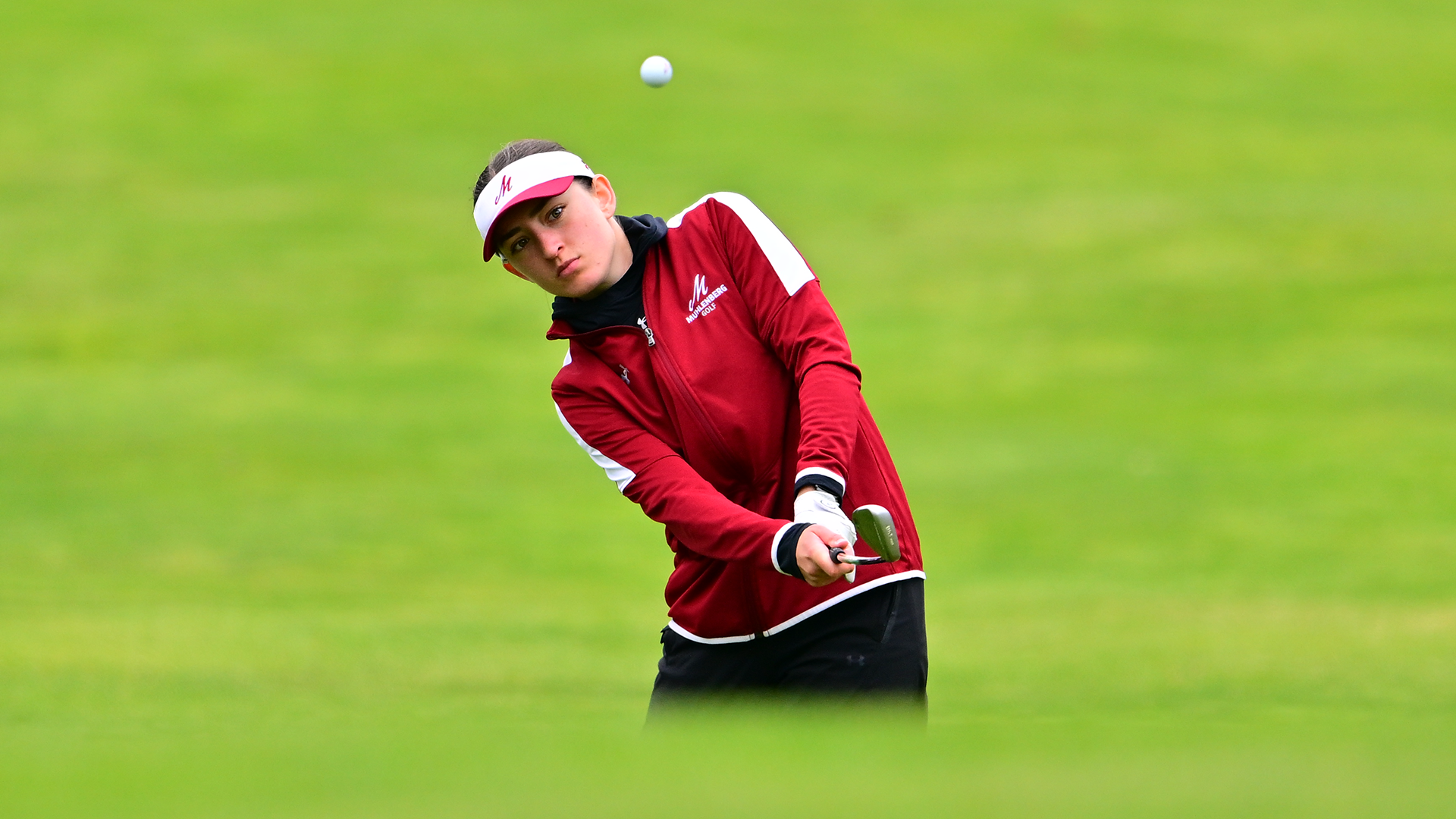 a women's golf in a red pullover and white visor chipping the ball