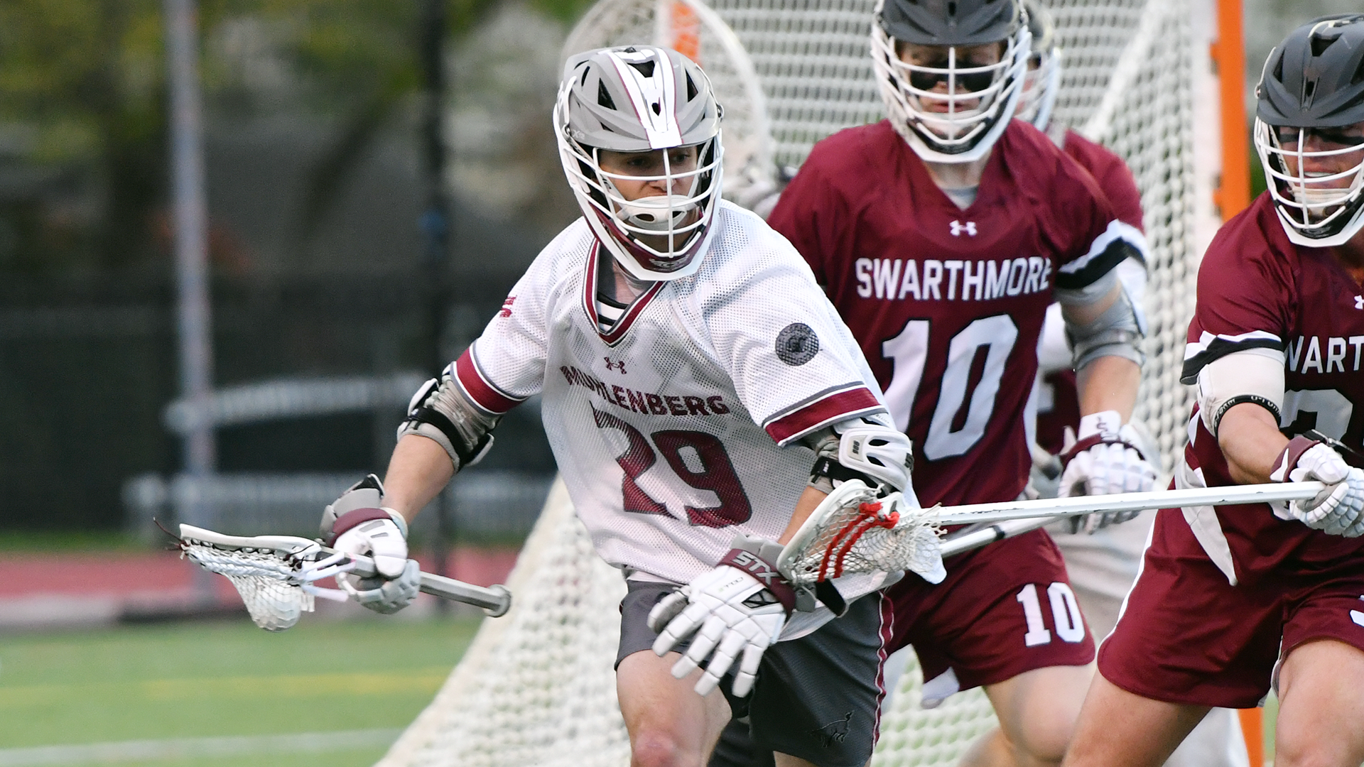 a men's lacrosse player in a white jersey and grey helmet cradling the ball with two players in red jersey defending him from behind