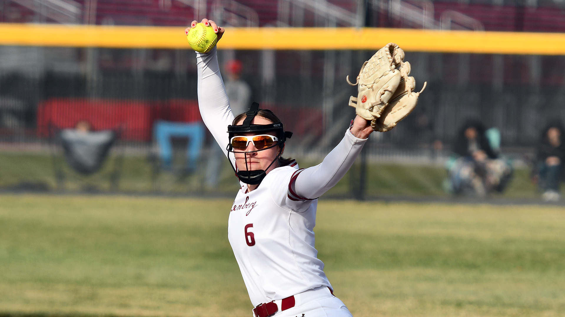a softball pitcher in a white jersey and white pants wearing number 6 brings her arm behind her head