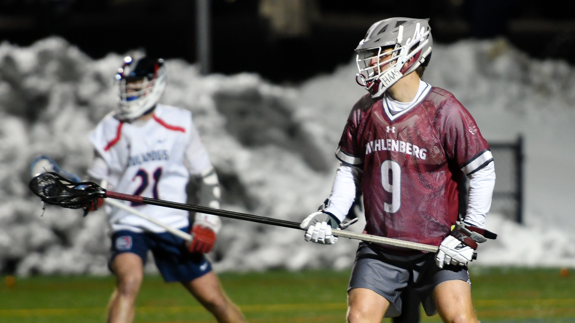 a men's lacrosse player with a long pole in a red uniform and grey helmet