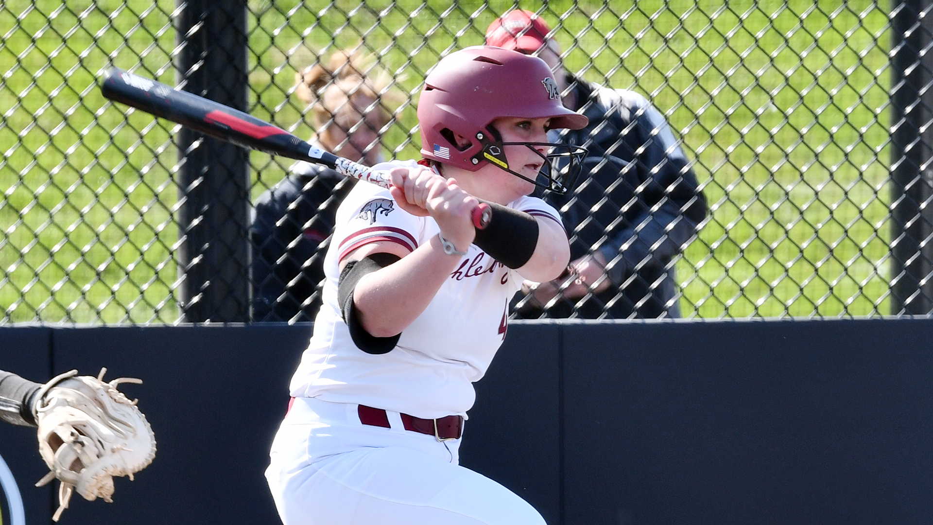 a left-handed softball batter in a white jersey and red helmet following through on her swing