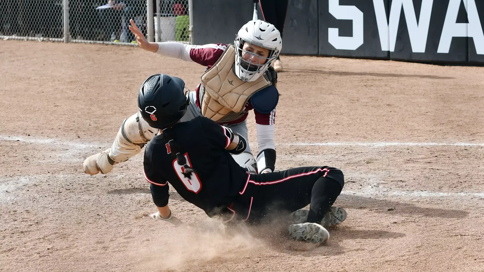 a softball catcher in a helmet and chest protector tagging out a sliding runner in a black uniform attempting to score
