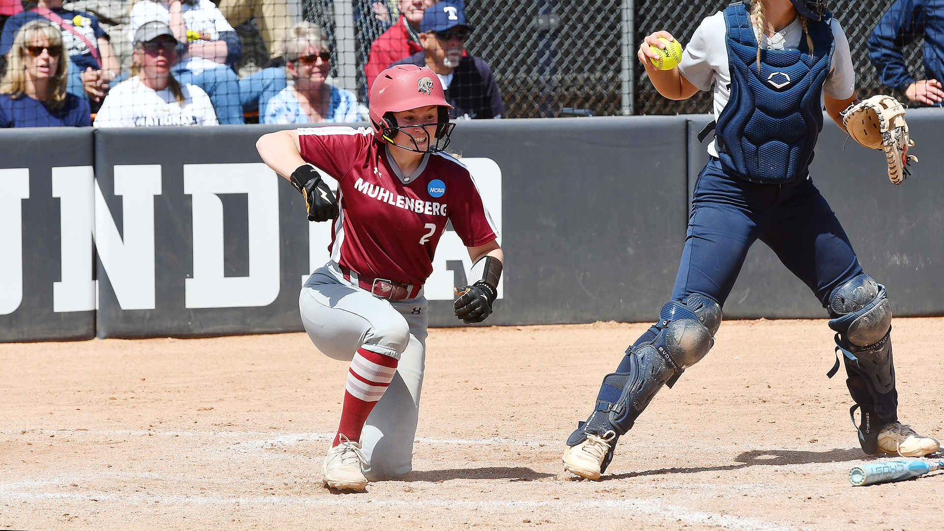 a softball player in a red jersey and red helmet celebrates after sliding in safely at home past a catcher dressed in blue