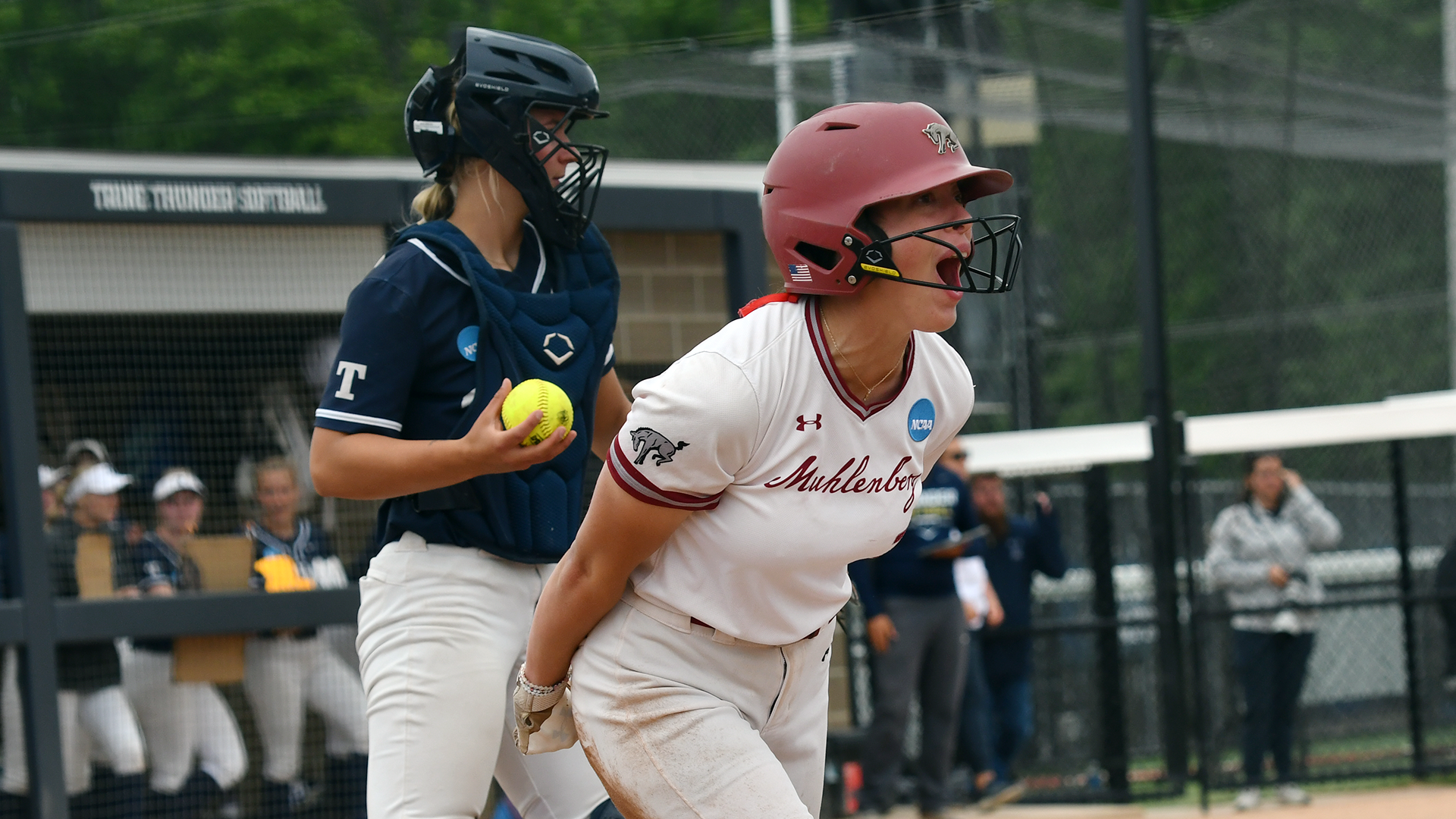 a softball player in a white jersey and red helmet yells while a catcher behind her in a blue mask and chest protector and white pants holds the ball 