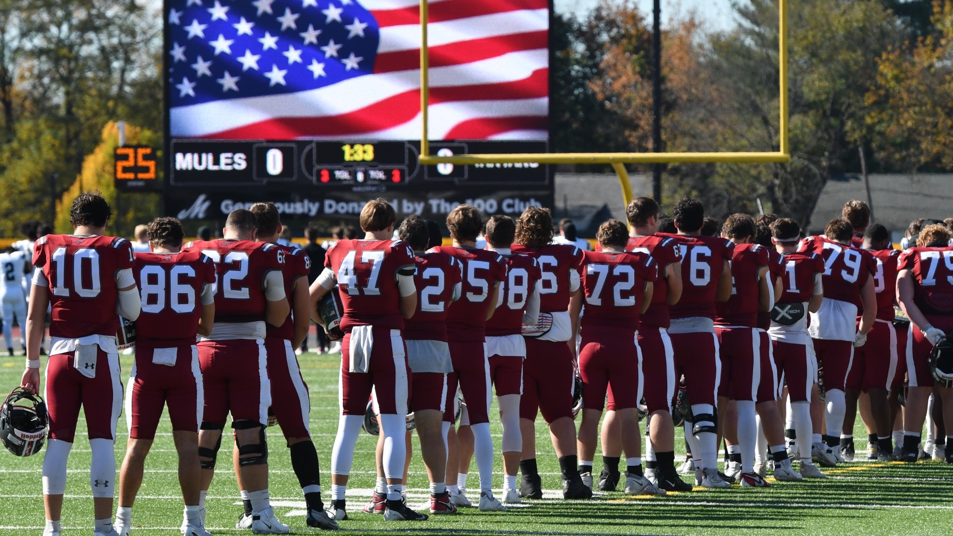 Football players in red uniforms stand along the sideline facing the field during a pregame ceremony as an American flag is displayed on the stadium scoreboard.