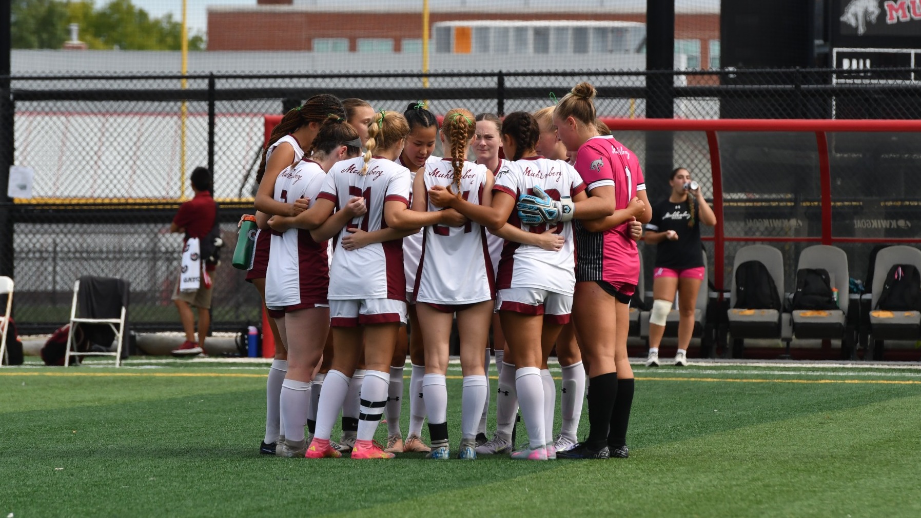 A women's soccer team in white uniforms huddles pregame in the middle of a field.