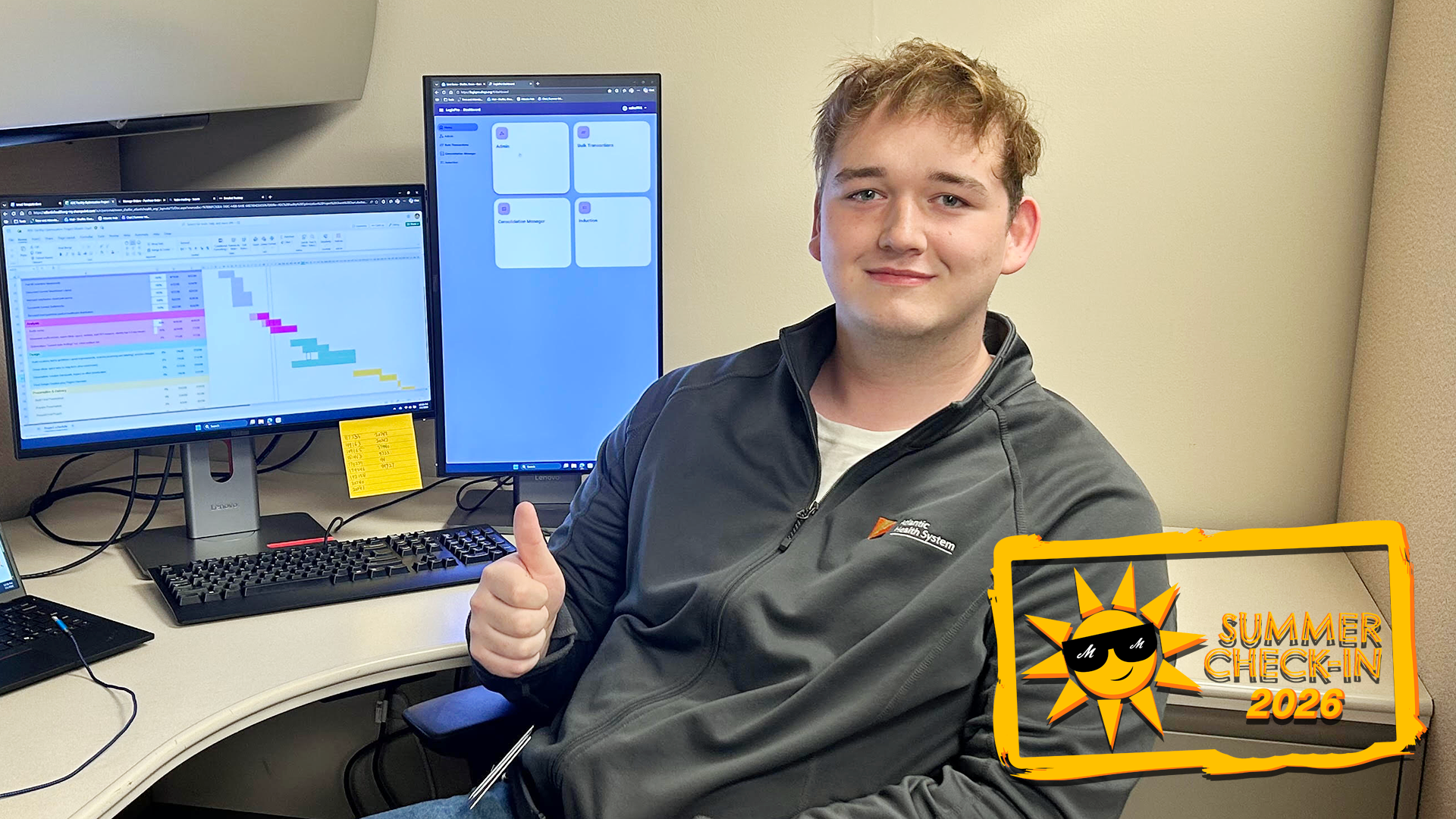 a young man in a grey zip-up top giving the thumbs-up sign while sitting at a workstation with two computer monitors