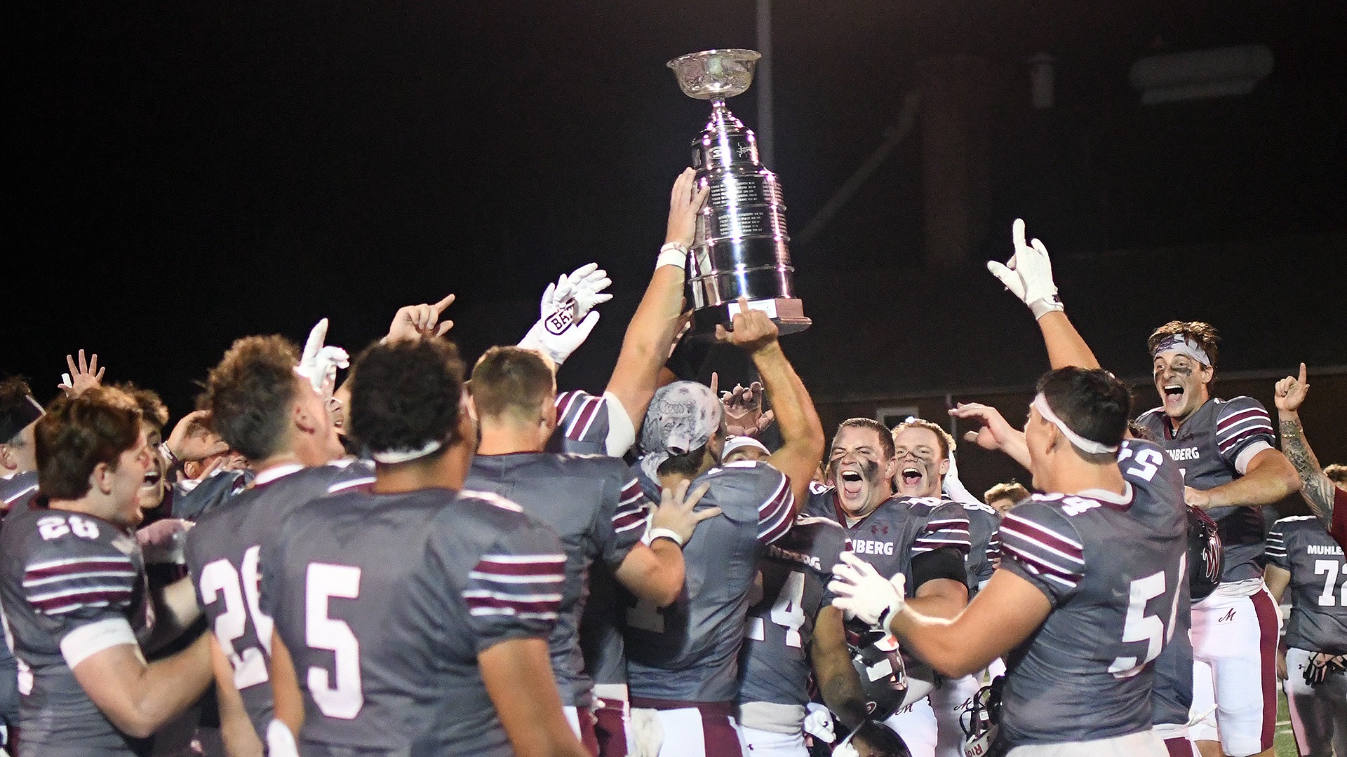 football players in grey uniforms raising a silver trophy after a night game