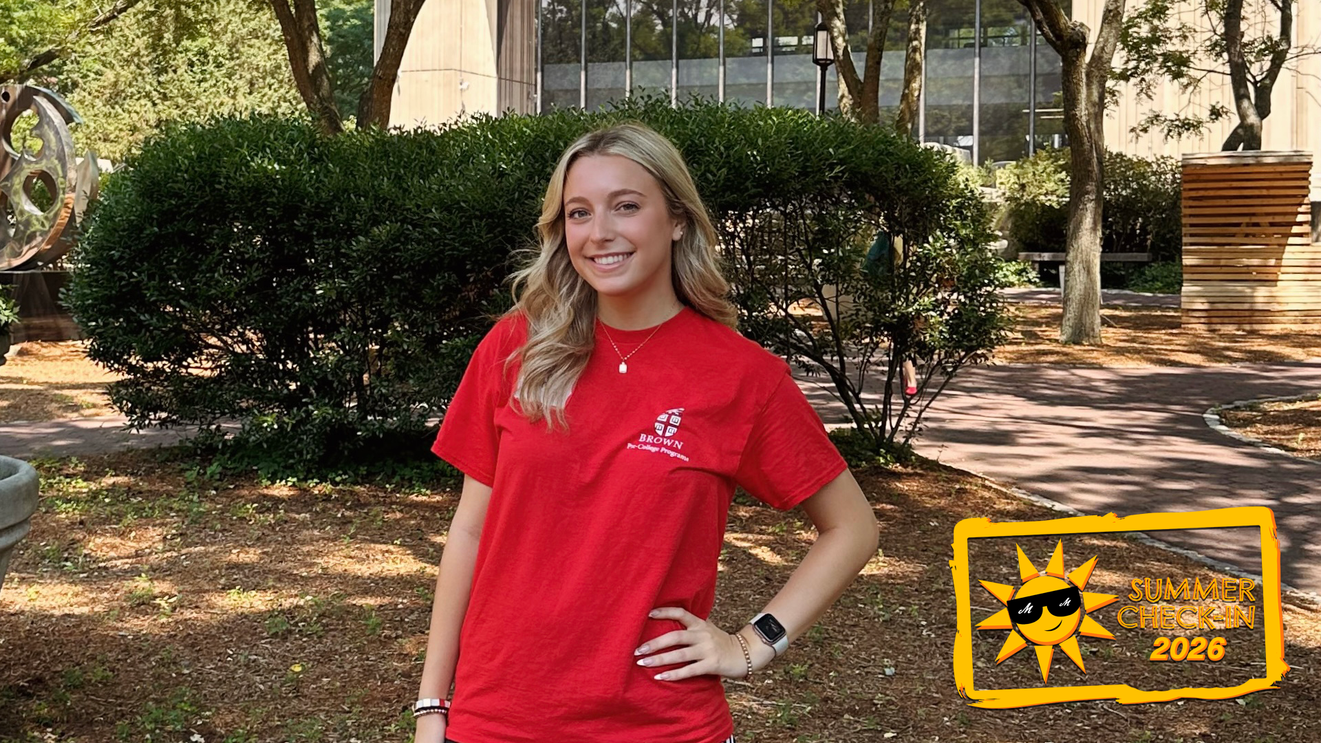 a young woman in a red t-shirt posing outdoors in front of a bush, with a building in the background