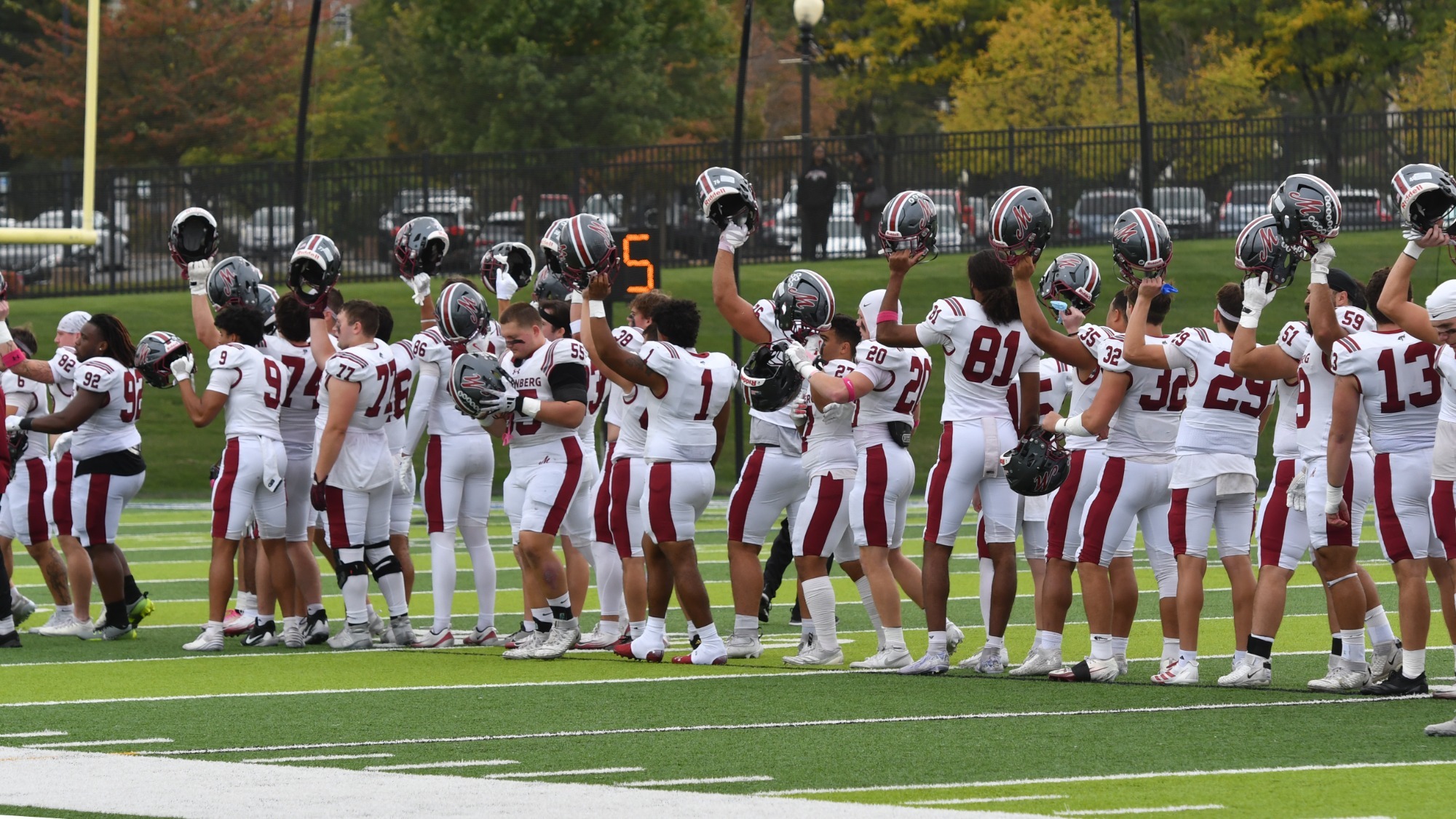 A group of football players in white uniforms holds their helmets up as part of a pregame ceremony