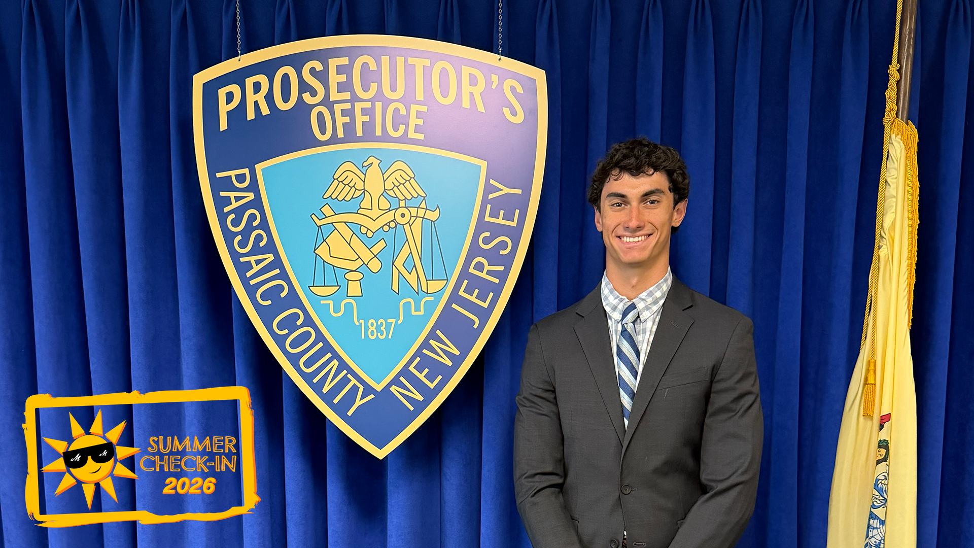 a young man in a suit and tie standing in front of a blue curtain on which a shield reading Prosecutor's Office, Passaic County, New Jersey is displayed