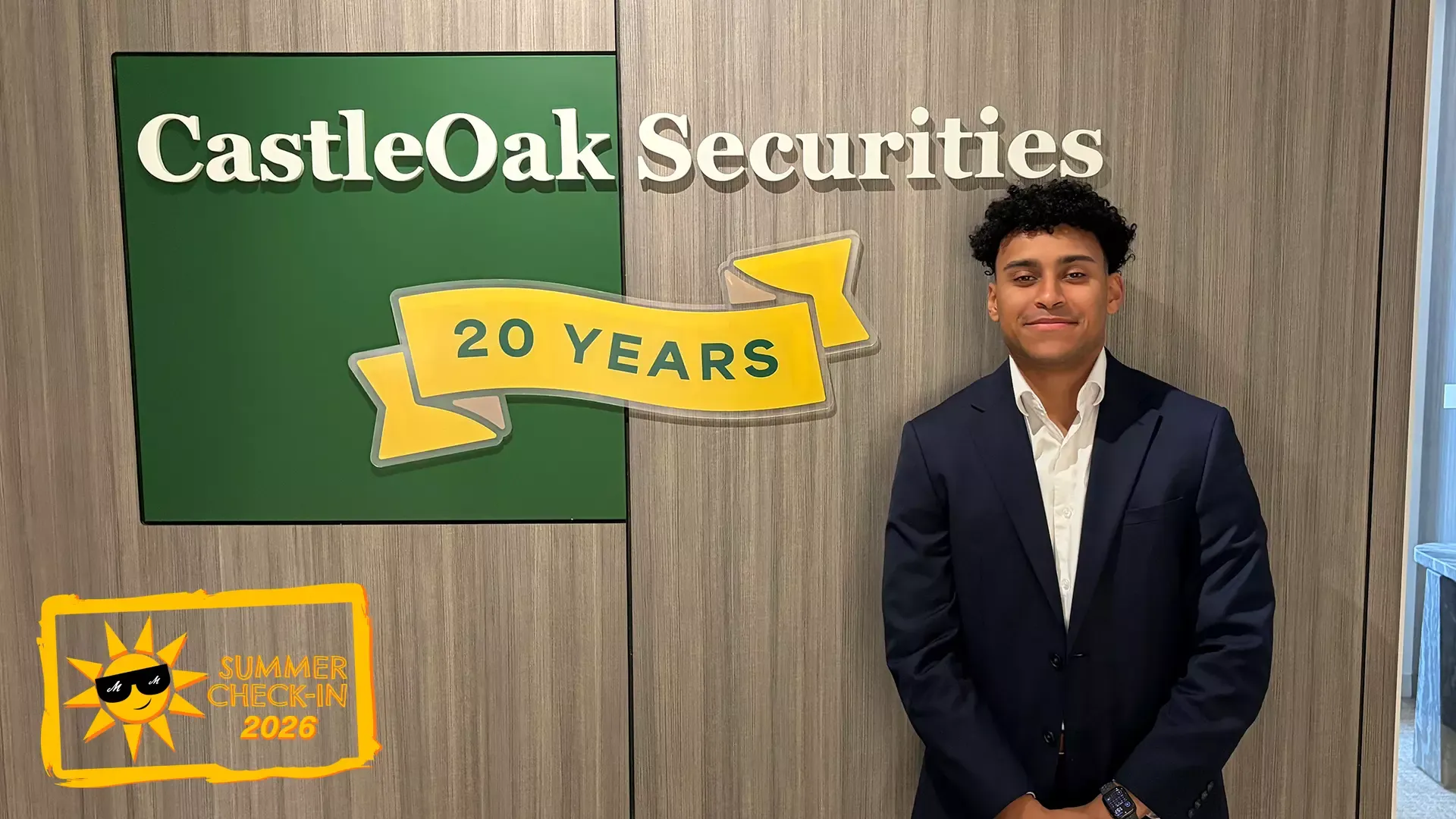 a young man in a blue suit and white shirt posing in front of a wall that features a green square with the words CastleOak Securities in white