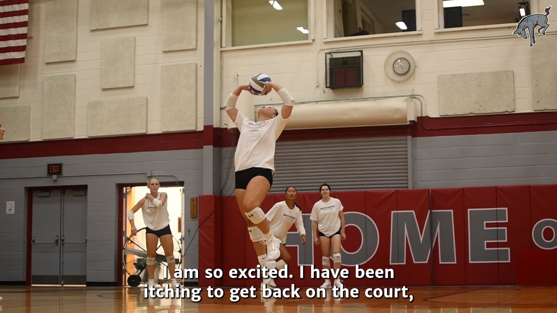 a volleyball player in a white t-shirt and black shorts setting a ball indoors with three teammates behind her