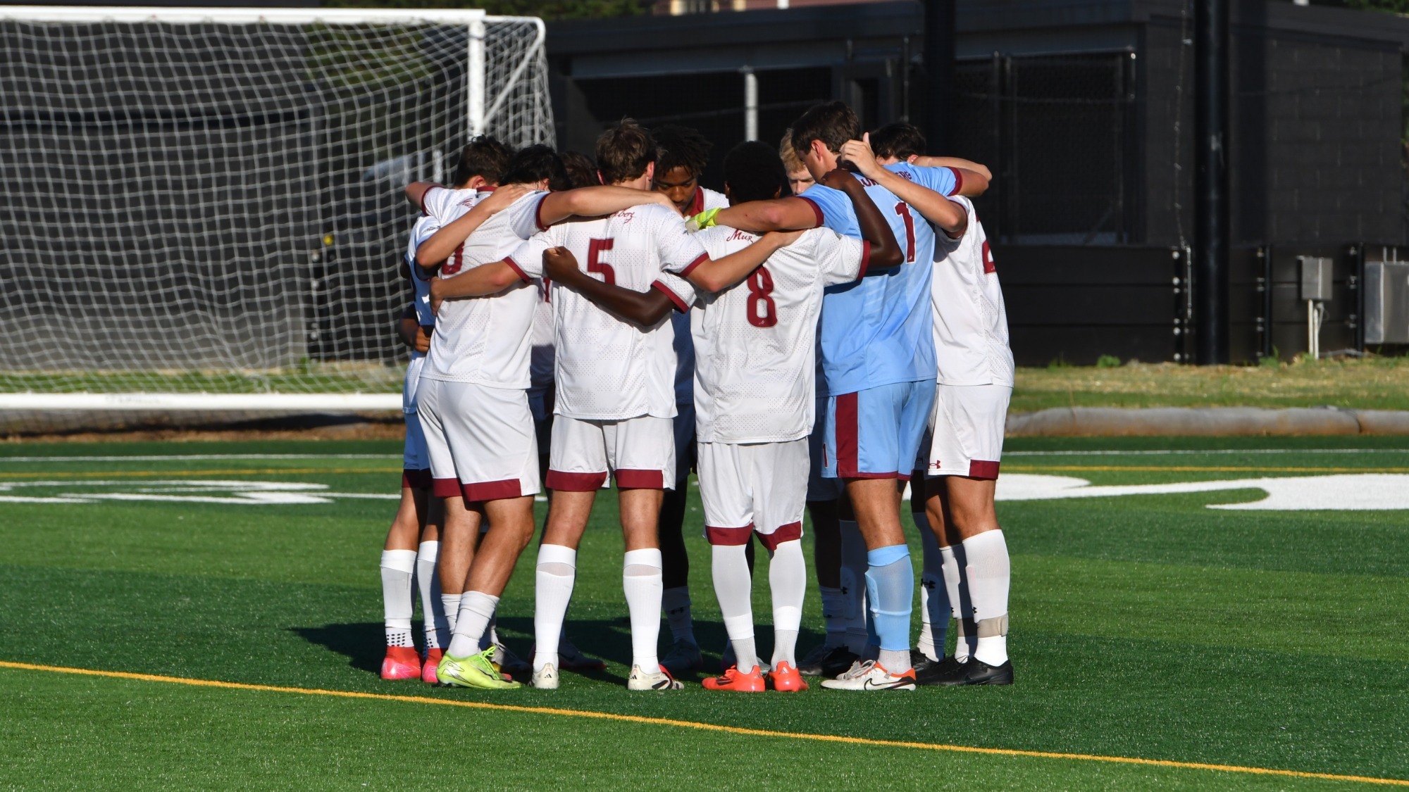 A group of men's soccer players clad in white except for the goalie huddle on the pitch
